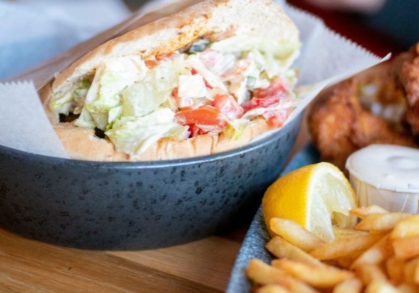 A sandwich with a mixed vegetable filling sits in a bowl next to a serving of fries, lemon wedges, fried chicken, and a small cup of sauce, representing the mindful consumption trend in modernizing lent.