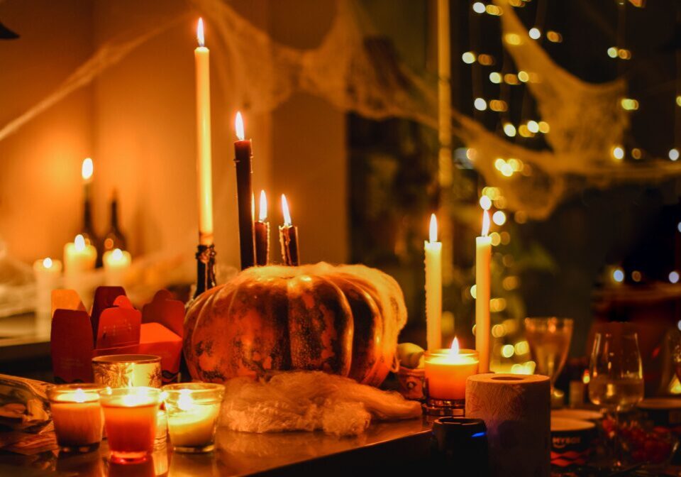 A dimly lit table decorated with lit candles, a large pumpkin, cobwebs, and string lights creates a warm, festive atmosphere perfect for Halloween.