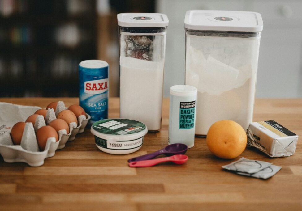 A variety of baking ingredients, including eggs, salt, sugar, flour, baking powder, mascarpone cheese, an orange, butter, and measuring spoons with labels to decode are arranged on a wooden countertop.