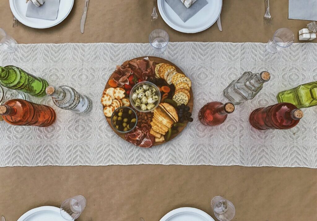 A charcuterie board with assorted meats, cheeses, crackers, and olives sits at the center of a table adorned with a lace runner, surrounded by glass bottles and plates set for a meal—a perfect display of 2023 food trends.