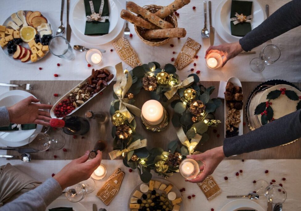Overhead view of a festive dining table setup with candles, a wreath centerpiece, assorted bread, cheese, fruits, nuts, and hands reaching for food and candles— perfect for stress-free entertaining when hosting for the holidays.