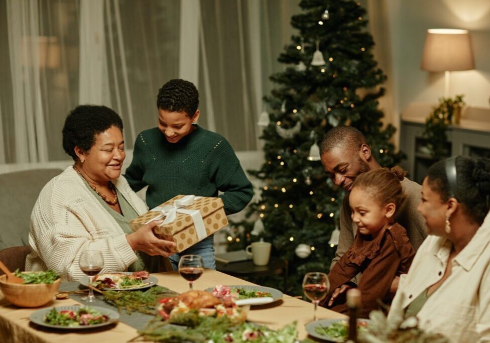 A group of people gather around a holiday table with food, as an older woman seated at the head of the table receives a gift from a young boy. A decorated Christmas tree is visible in the background, highlighting the joys of hosting for the holidays and stress-free entertaining.
