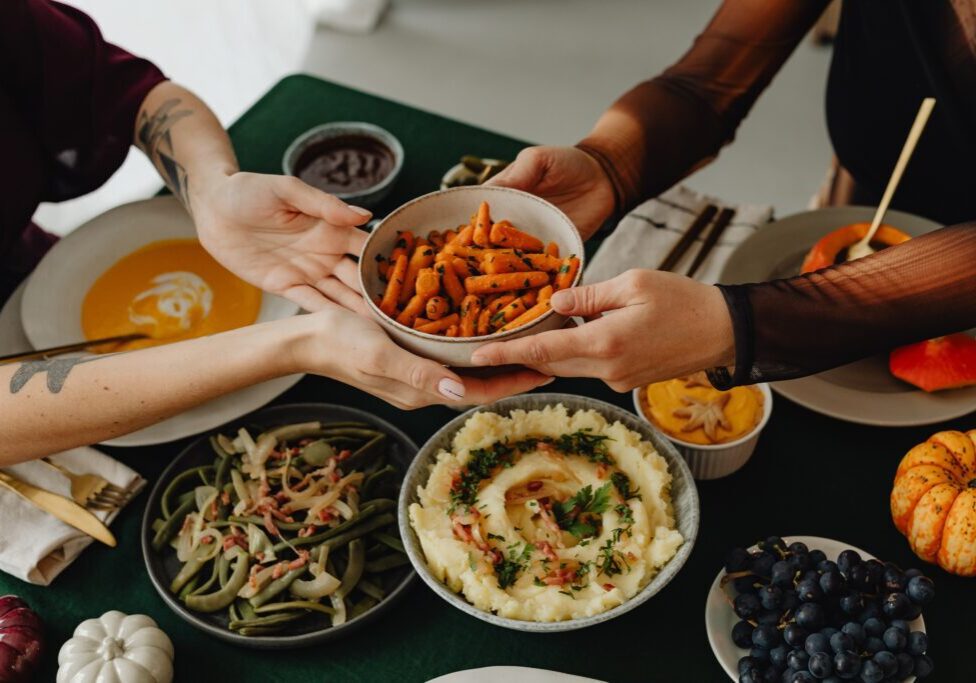 Two friends at a Friendsgiving dinner pass a bowl of roasted carrots across a table set with various dishes, including mashed potatoes, green beans, grapes, and soup.