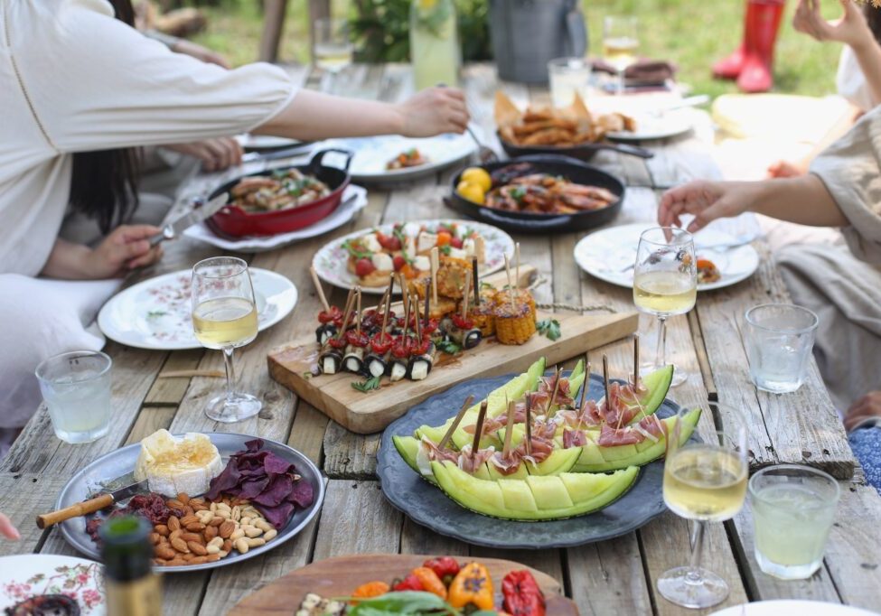 A group of people seated around a wooden picnic table covered with various dishes, such as fruit, skewers, cheese, and wine glasses with white wine, enjoying a meal together outdoors. The scene captures the essence of simple hospitality as they savor the joys of in-home entertaining.