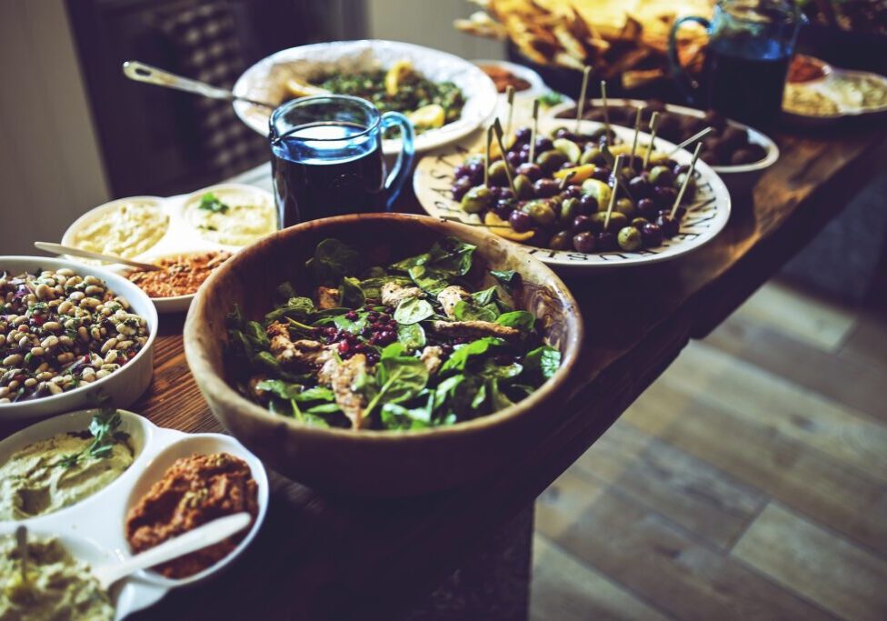 A table is set with various dishes, including a large bowl of salad rich in antioxidants, skewered olives, small bowls of dips, and plates of assorted vegetables and snacks.