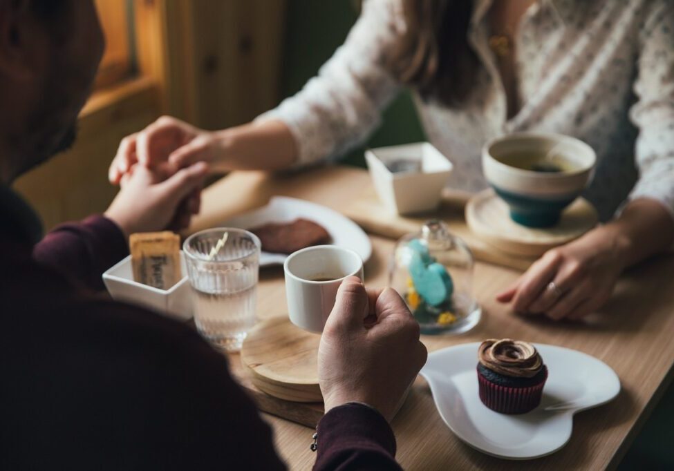 Two people sit at a table holding hands, with assorted desserts and beverages in front of them, reminiscing about their fun cooking class earlier that day.