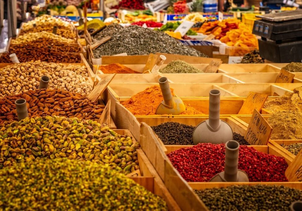 Market stall featuring wooden crates filled with various spices, herbs, nuts, and dried fruits in vibrant colors, organized with scoops and price tags. Discover fall spices from around the world that go beyond pumpkin spice to elevate your seasonal dishes.