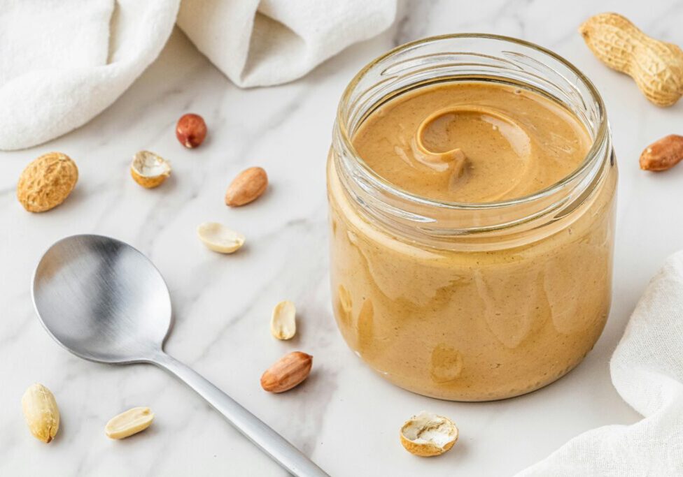 An open jar of peanut butter on a marble surface, surrounded by peanuts in shells and a metal spoon.