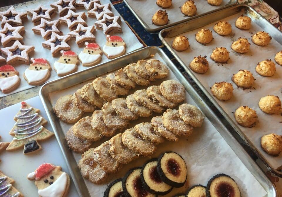 Various types of holiday-themed cookies, including star-shaped, Santa faces, tree-shaped, round sugar-coated, and round cookies with jam centers, are arranged on baking trays. The delightful assortment showcases the joy of holiday baking at its finest.