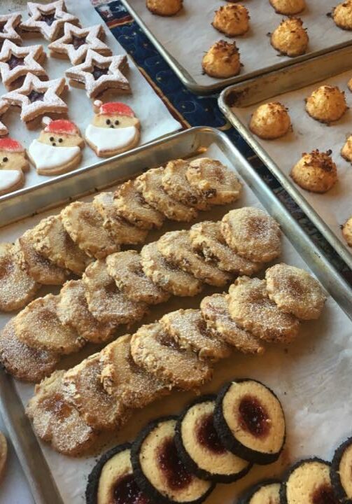 Various types of holiday-themed cookies, including star-shaped, Santa faces, tree-shaped, round sugar-coated, and round cookies with jam centers, are arranged on baking trays. The delightful assortment showcases the joy of holiday baking at its finest.